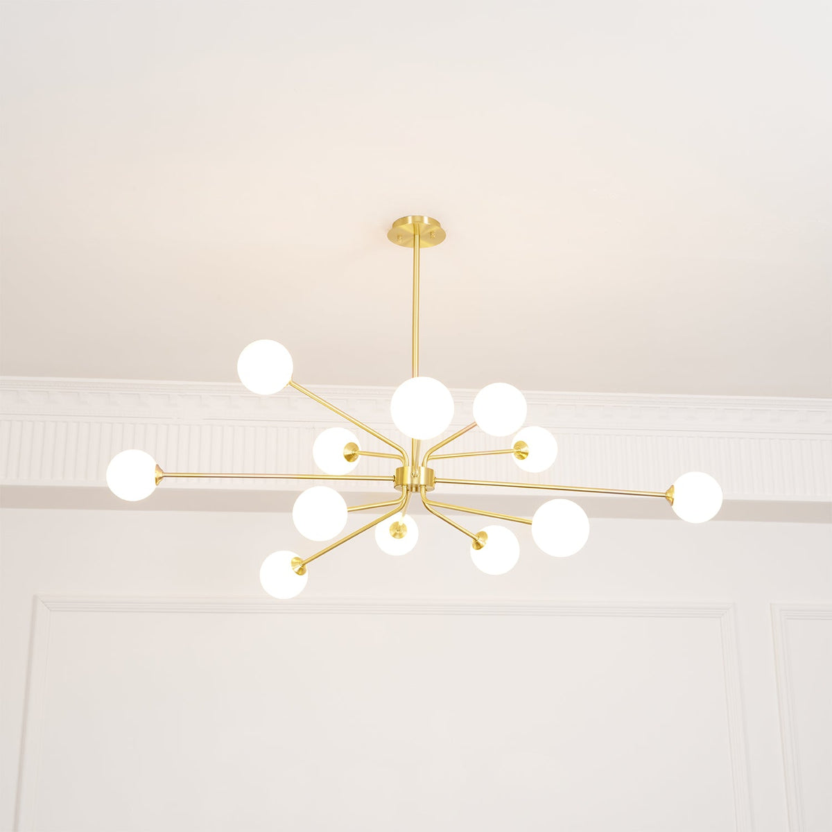 Brass Clérachic chandelier with glowing white globes on long arms, seen below a white ceiling with moulded panels.