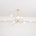 A brass Clérachic chandelier with a wide spread of arms and white globes, lit against a white ceiling.