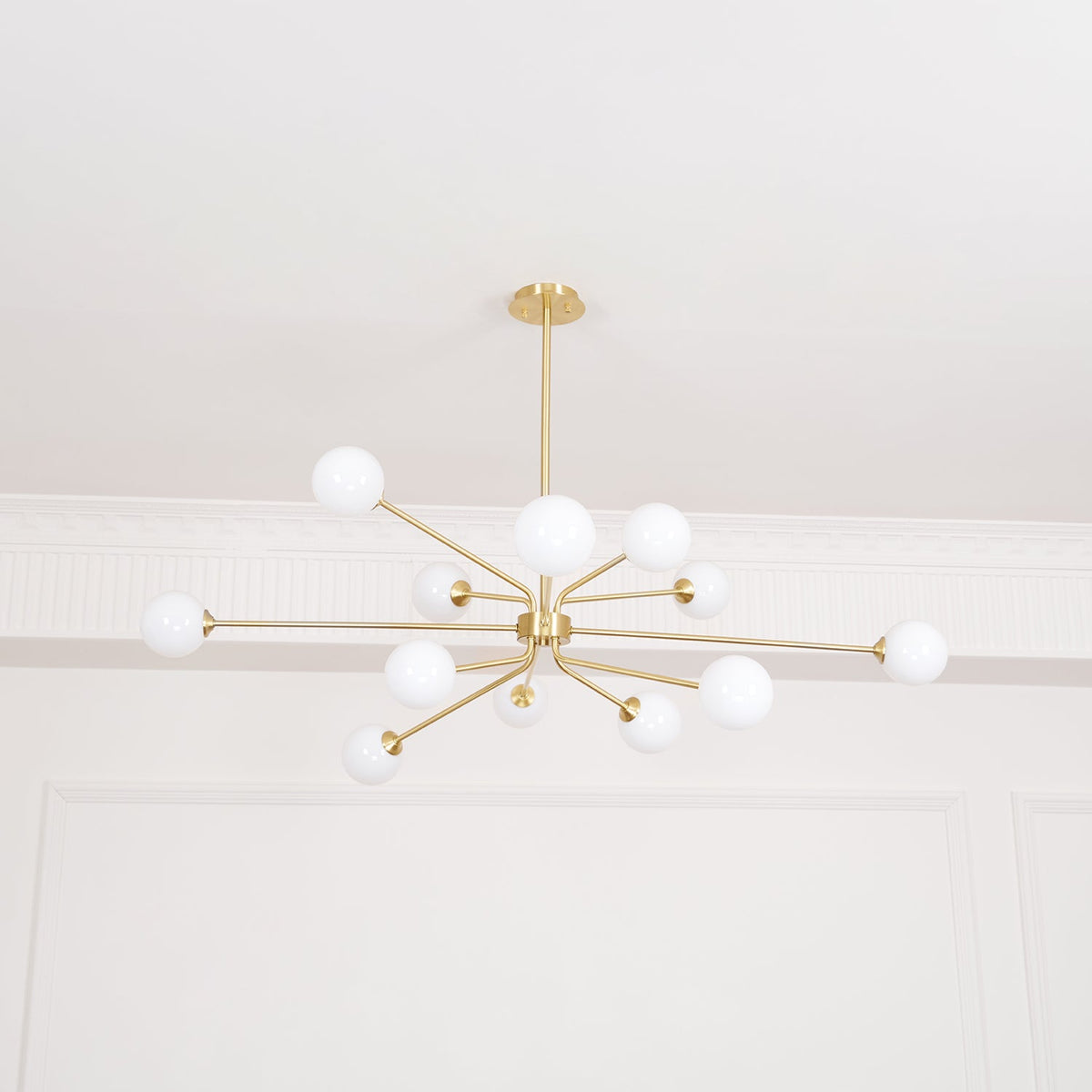 A brass Clérachic chandelier with a wide spread of arms and white globes, lit against a white ceiling.