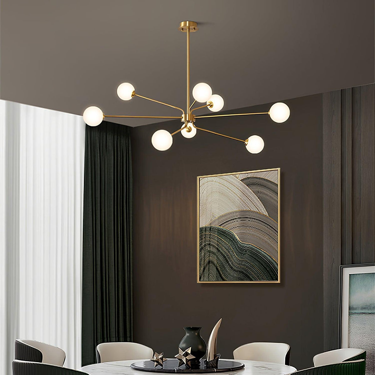 A brass Clérachic chandelier with white globes hangs in a dining room with a round white table, green curtains and abstract art.