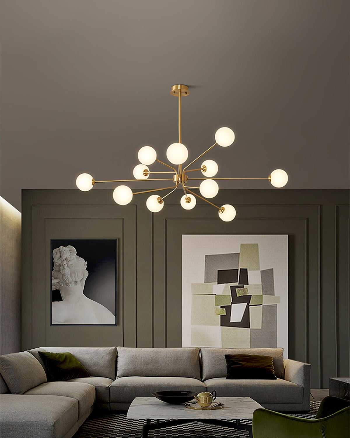 Wide view of a lit brass Clérachic chandelier above a sofa and coffee table, with two framed prints on olive panelling.