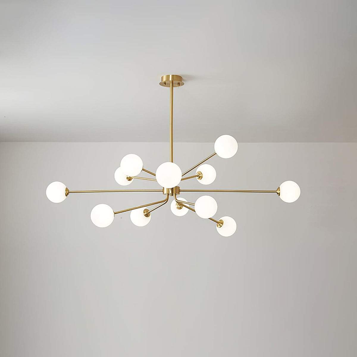 Large brass Clérachic chandelier with glowing white globes on many arms, hanging under a light grey ceiling.
