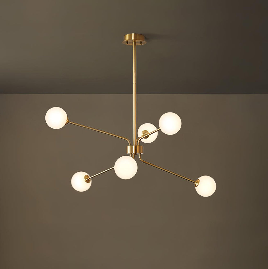 A smaller brass Clérachic chandelier with six white globes hangs from a long rod in front of a dark olive wall.