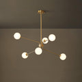 A smaller brass Clérachic chandelier with six white globes hangs from a long rod in front of a dark olive wall.