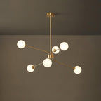 A smaller brass Clérachic chandelier with six white globes hangs from a long rod in front of a dark olive wall.