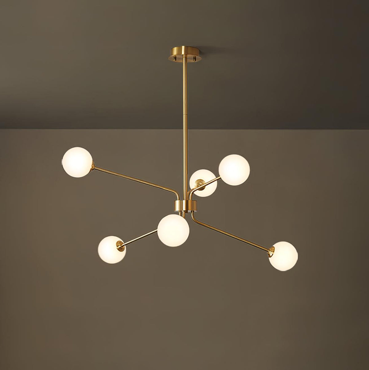 A smaller brass Clérachic chandelier with six white globes hangs from a long rod in front of a dark olive wall.