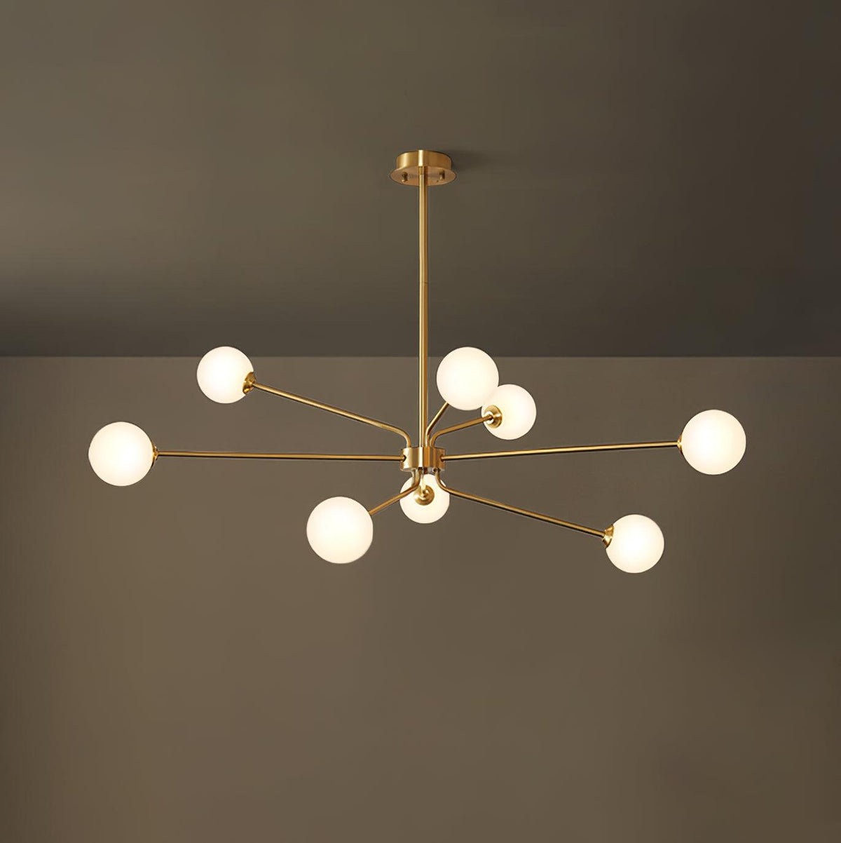 Brass Clérachic chandelier with nine glowing white globes on long arms, in front of a dark olive wall.