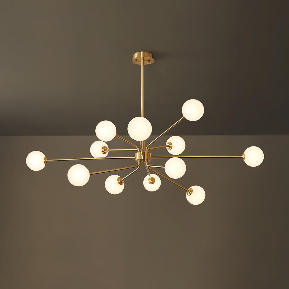 A large brass Clérachic chandelier with a full cluster of glowing white globes, against a dark olive wall and ceiling.