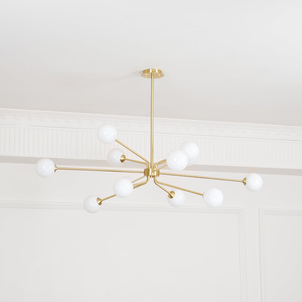 Lit brass Clérachic chandelier with white globes on slim arms, below a white ceiling with decorative mouldings.