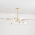Lit brass Clérachic chandelier with white globes on slim arms, below a white ceiling with decorative mouldings.