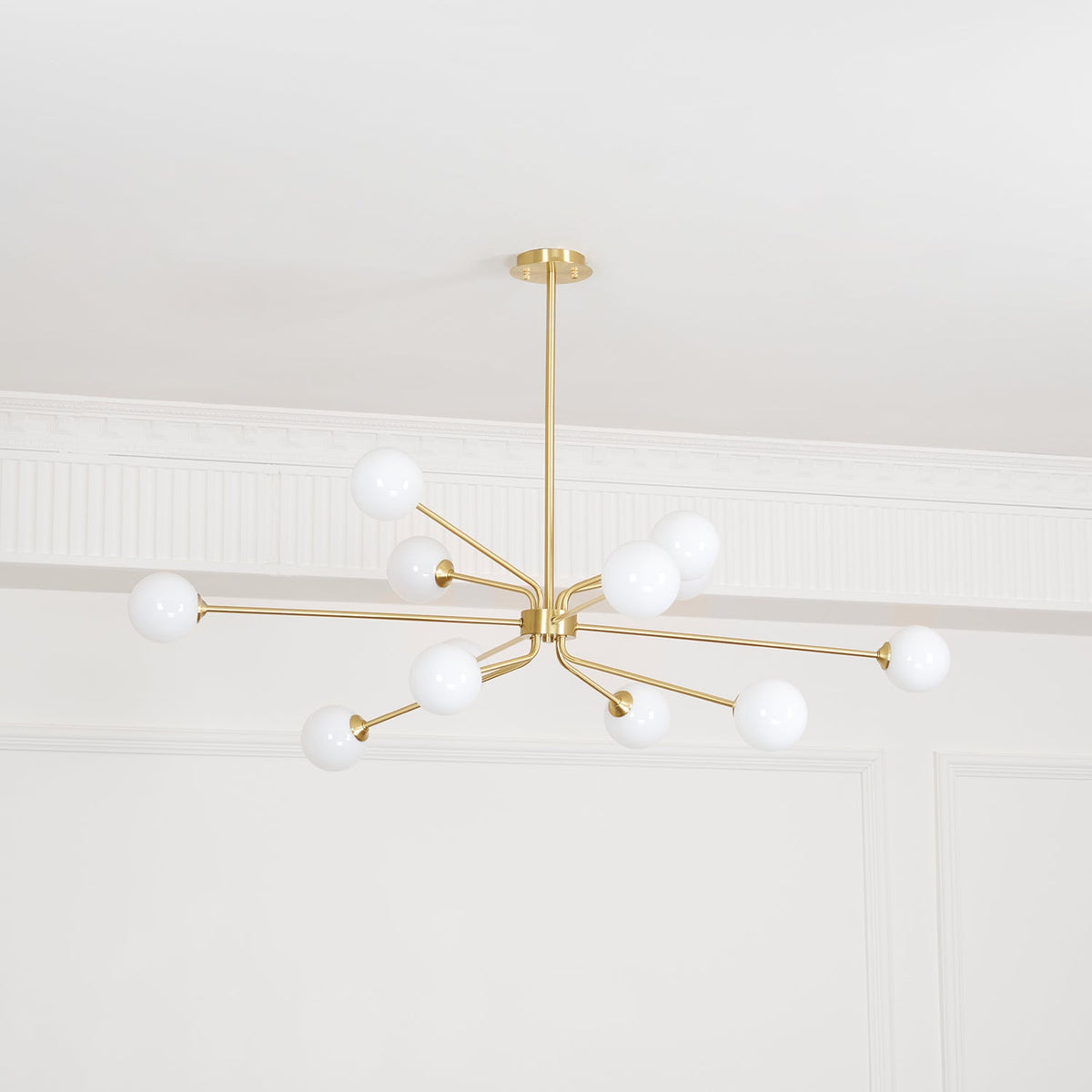 Lit brass Clérachic chandelier with white globes on slim arms, below a white ceiling with decorative mouldings.