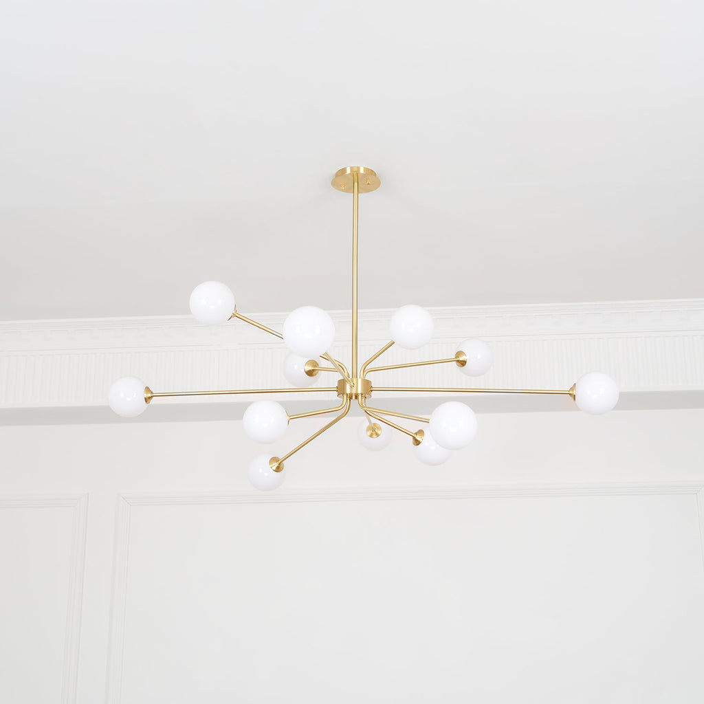 A brass Clérachic chandelier with many arms and white globes hangs below a plain white ceiling, seen from far below.