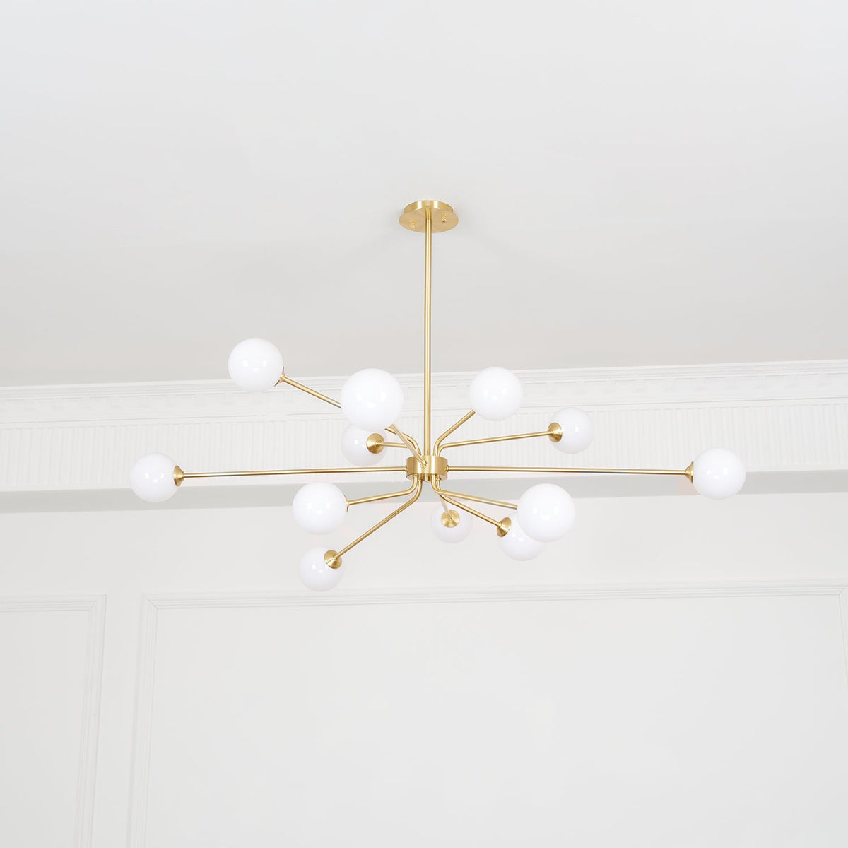 A brass Clérachic chandelier with many arms and white globes hangs below a plain white ceiling, seen from far below.