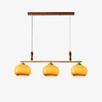 This Clérachic three-head bar pendant on a plain light backdrop, with amber glass shades, a brass bar and walnut-toned wooden accents.