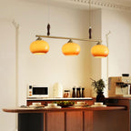 Three amber glass shades on a brass bar, a Clérachic pendant hanging over a kitchen counter with a coffee machine and warm timber cabinets.