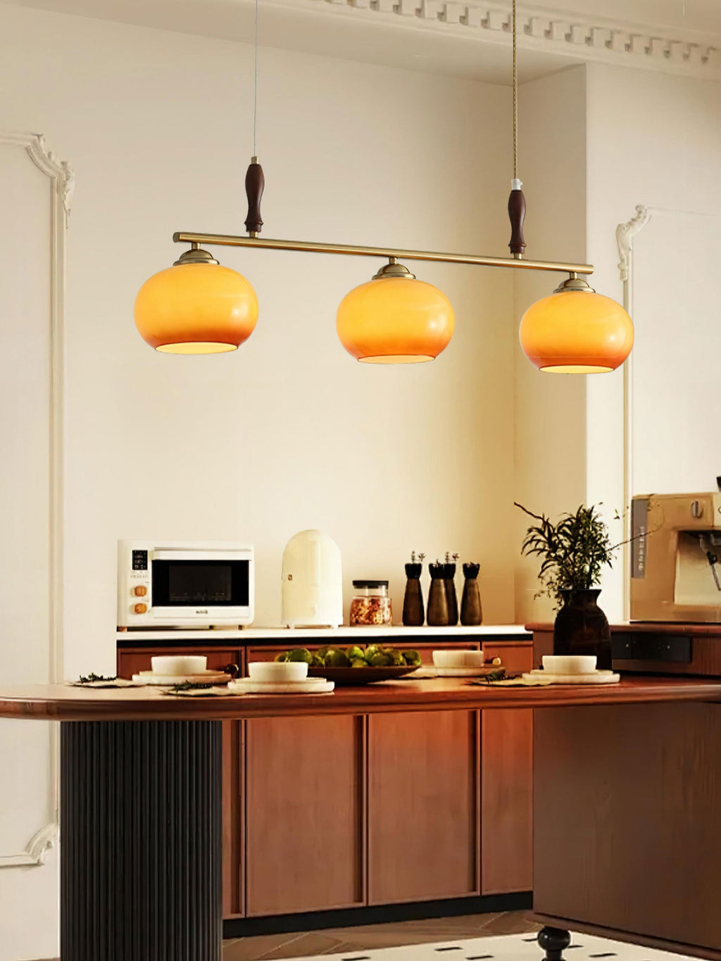 Clérachic bar pendant with three glowing amber shades above a kitchen island, with a microwave, spice mills and a plant behind.