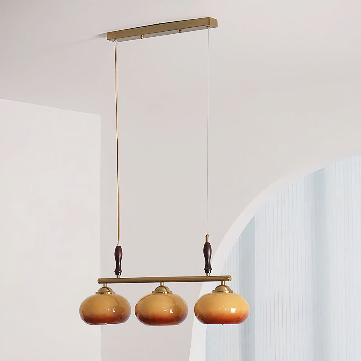 Low view of a Clérachic three-head pendant hanging from a slim brass ceiling plate beside a white archway, its amber shades unlit.