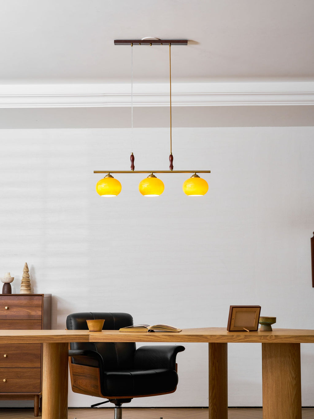 A Clérachic bar pendant with three bright yellow-lit shades, hanging over a wooden desk with a black chair in a room with a white panelled wall.