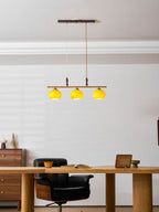 A Clérachic bar pendant with three bright yellow-lit shades, hanging over a wooden desk with a black chair in a room with a white panelled wall.