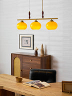 Lit amber shades of a Clérachic pendant over a pale wood desk, with a framed print and a walnut sideboard with a cane door behind.