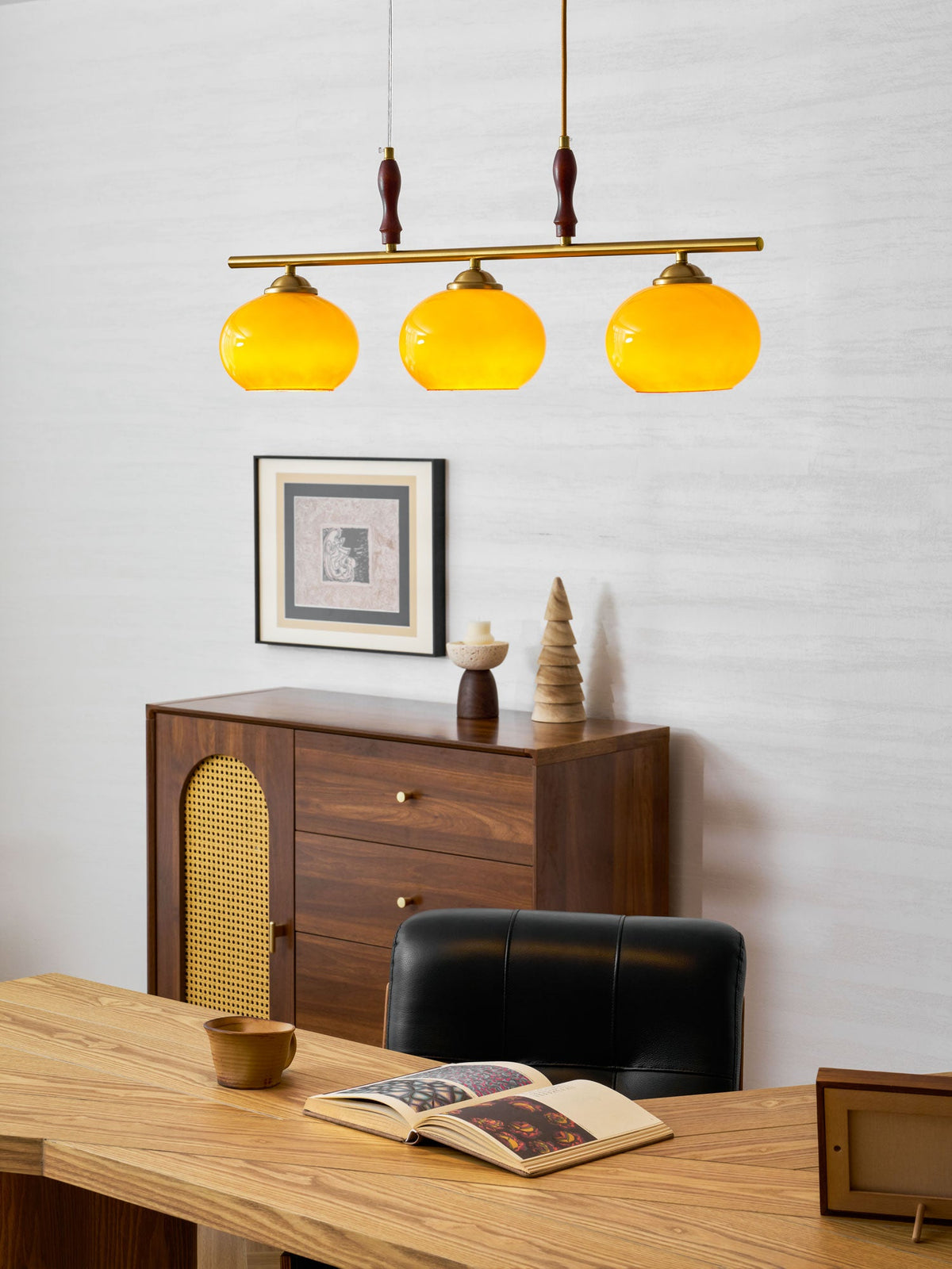 Lit amber shades of a Clérachic pendant over a pale wood desk, with a framed print and a walnut sideboard with a cane door behind.