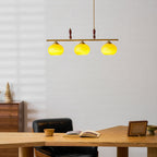 Clérachic pendant with three brightly lit shades glowing yellow-green, above a desk with an open book and a walnut chest of drawers.