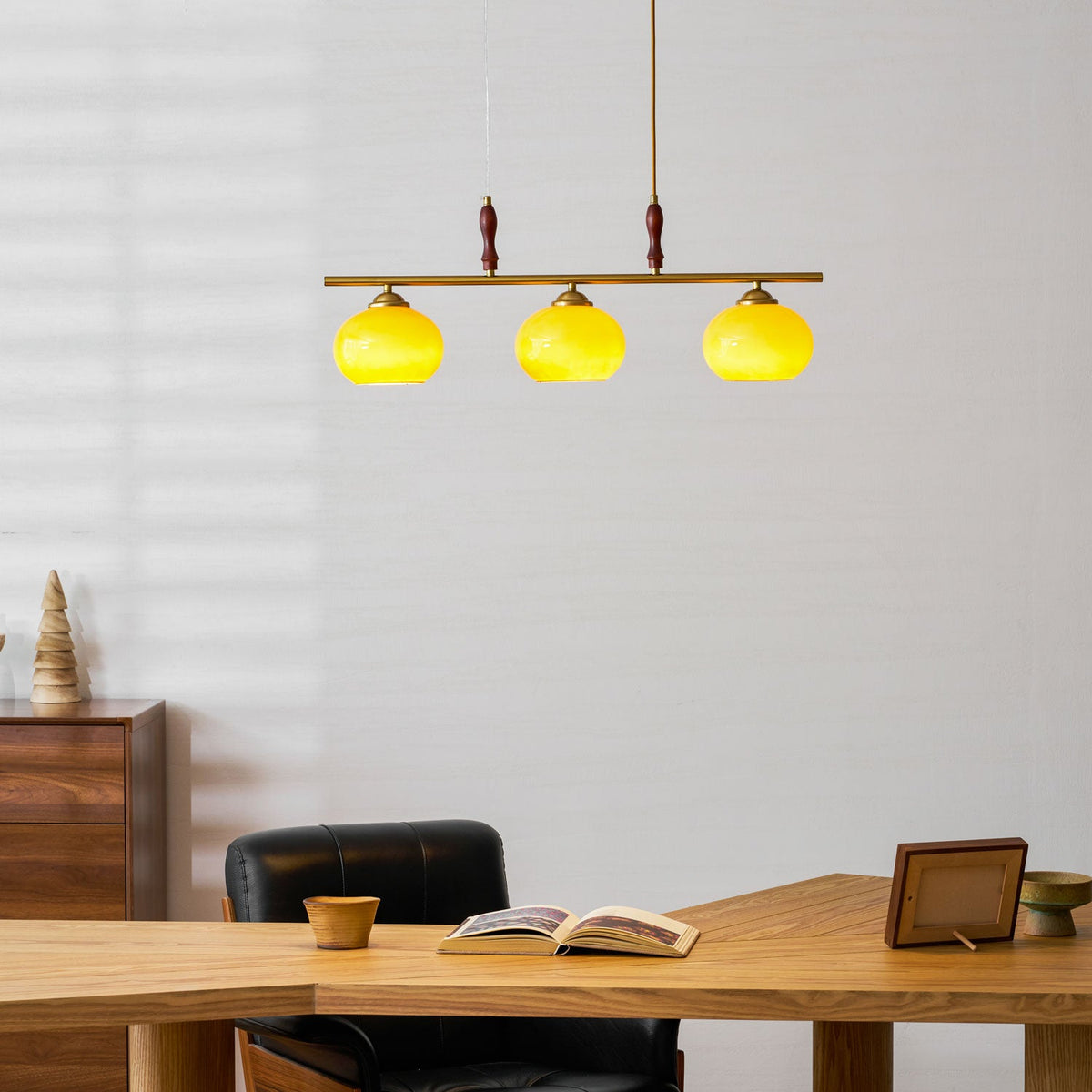 Clérachic pendant with three brightly lit shades glowing yellow-green, above a desk with an open book and a walnut chest of drawers.