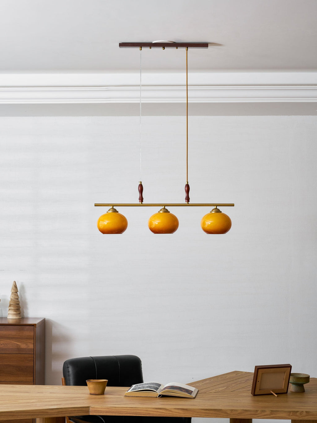 Small, distant view of a Clérachic three-shade pendant above a desk, with a black chair and a framed photograph at the table's edge.