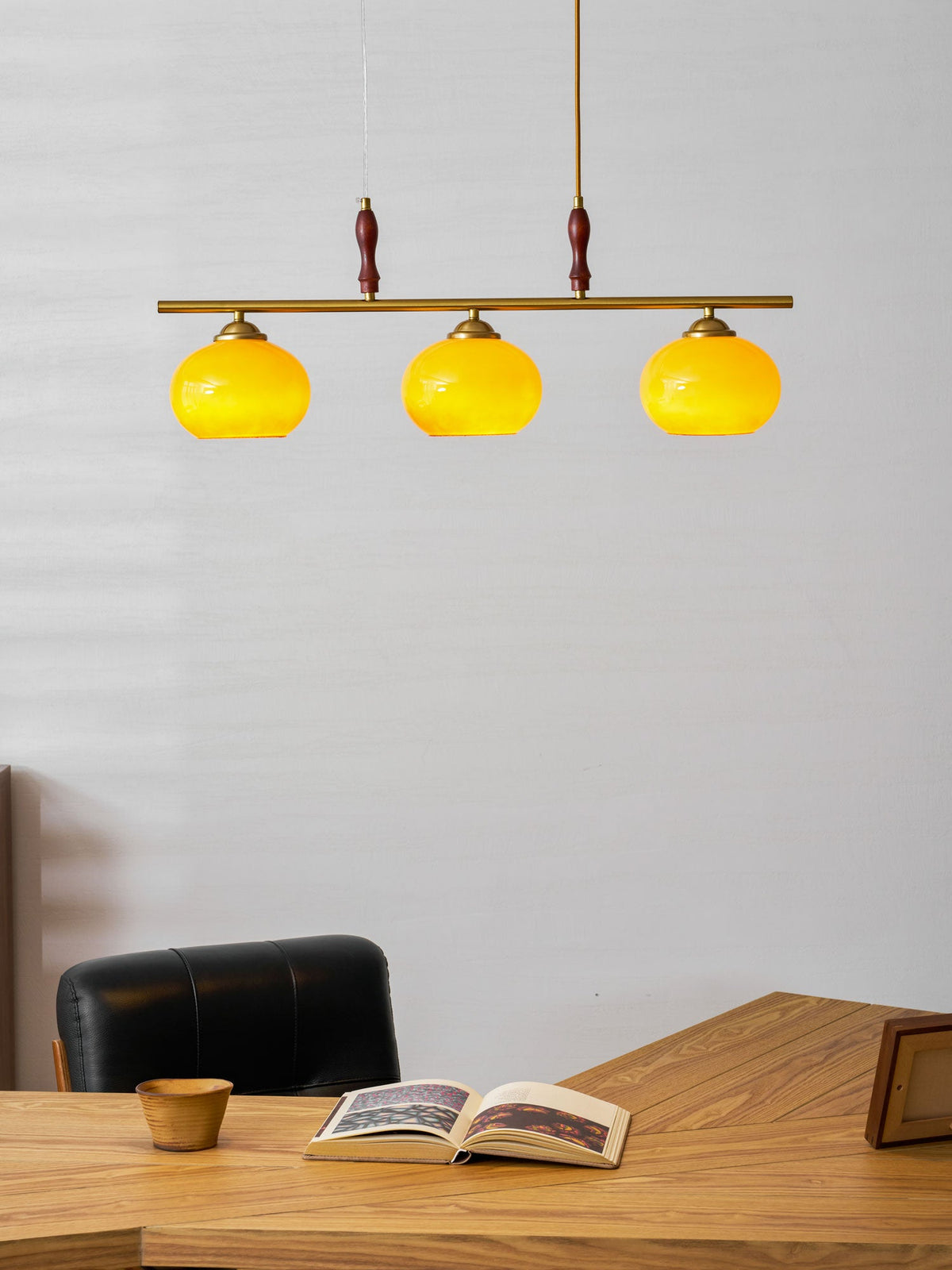 Close, low view of the Clérachic pendant's three glowing amber shades on a brass bar, above a desk with a wooden cup and an open book.