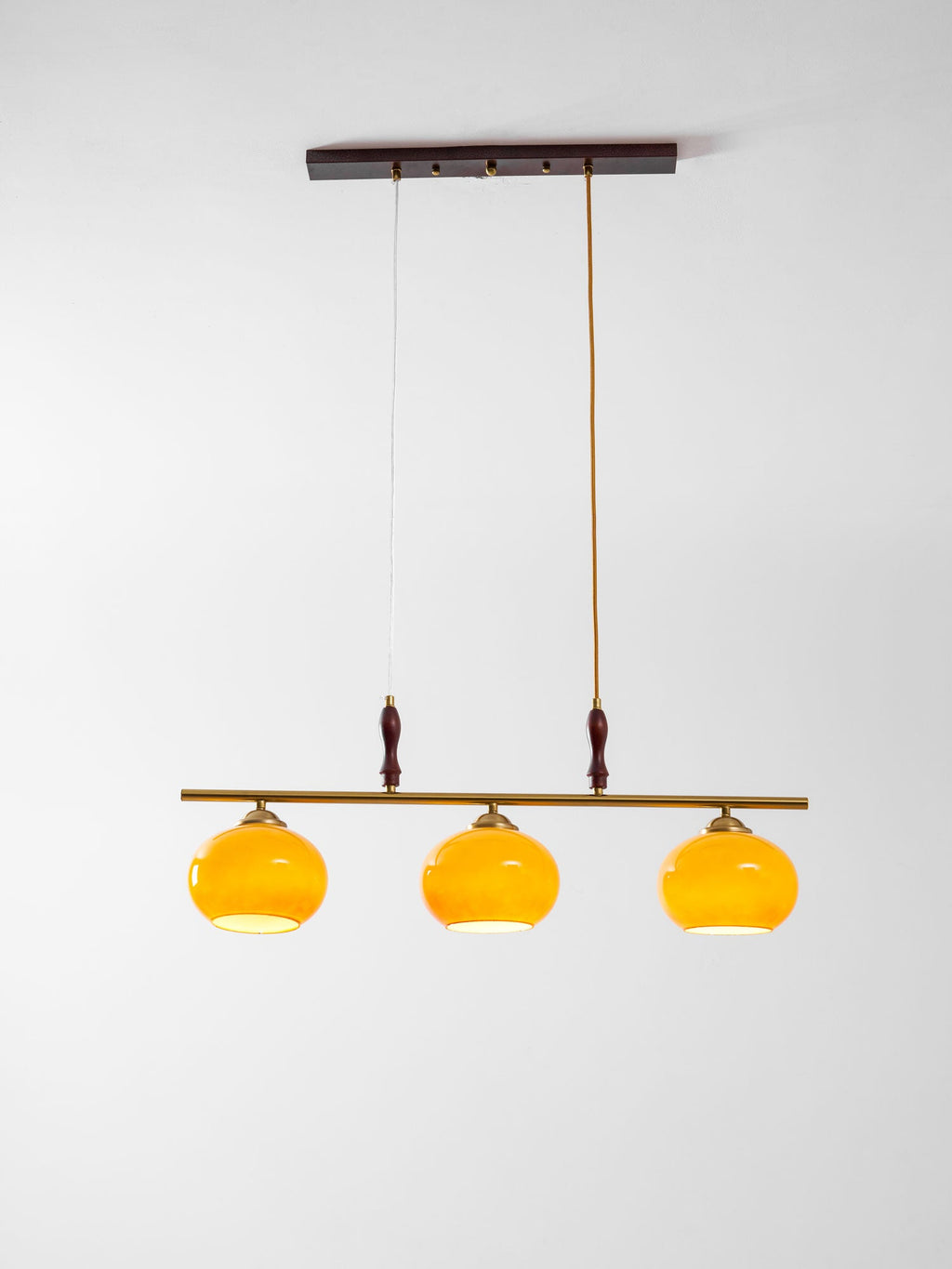 Clérachic pendant with three amber shades lit from within, hanging below a dark wooden ceiling plate against a soft grey backdrop.