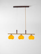 Clérachic pendant with three amber shades lit from within, hanging below a dark wooden ceiling plate against a soft grey backdrop.