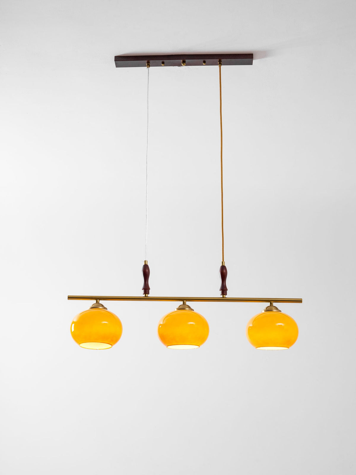 Clérachic pendant with three amber shades lit from within, hanging below a dark wooden ceiling plate against a soft grey backdrop.