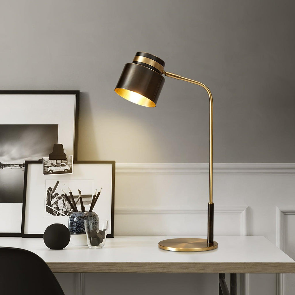 A Clérachic table lamp glowing on a white desk beside framed prints, its black shade lined in gold and lit against a grey wall.