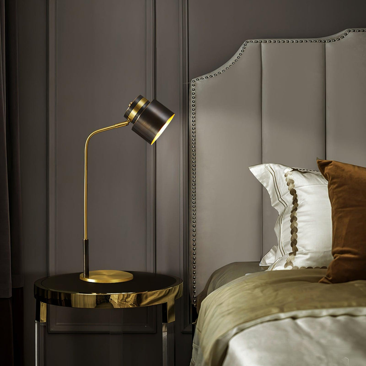 A Clérachic table lamp on a round bedside table with a polished metal edge, beside a grey headboard and olive bedding.