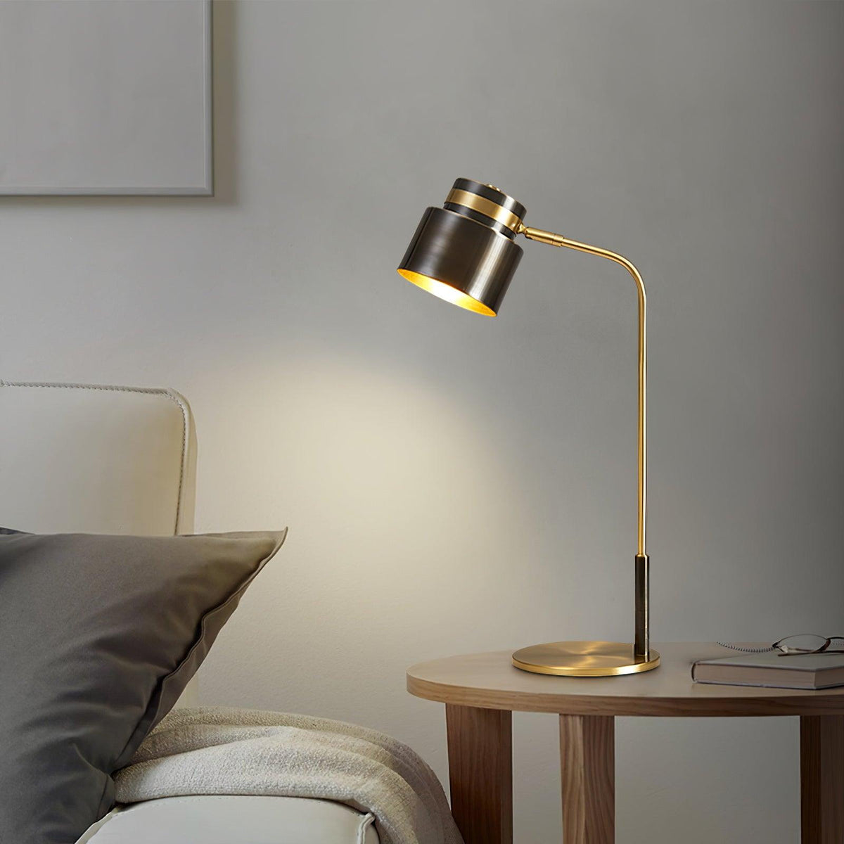 Lit softly beside a bed, a Clérachic table lamp rests on a round wooden table with a pair of glasses and a book.