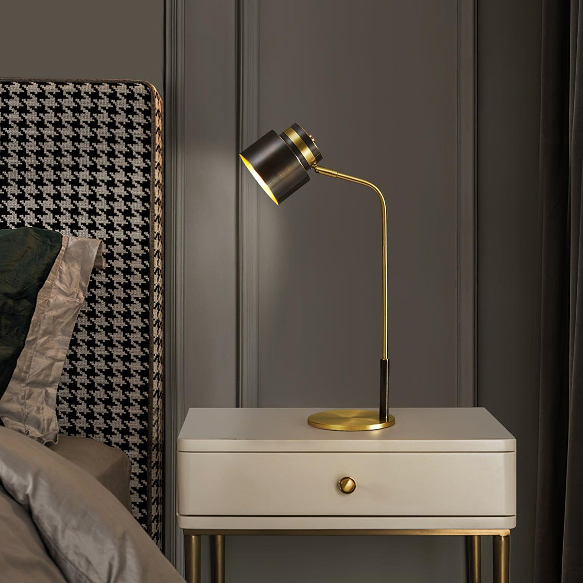 A Clérachic lamp on a cream bedside cabinet with a single drawer, next to a houndstooth headboard in a dark grey room.