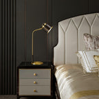 The Clérachic lamp on a black bedside chest with grey drawers, in front of a cream headboard and champagne bedding.