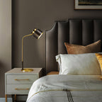 A Clérachic table lamp on a pale cabinet with brass pulls, with a padded taupe headboard and layered ivory bedding.