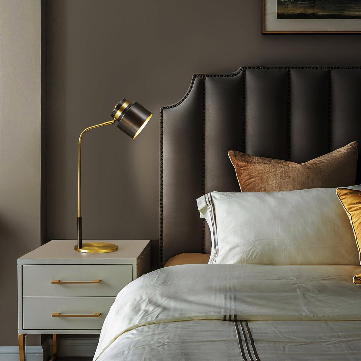 A Clérachic table lamp on a pale cabinet with brass pulls, with a padded taupe headboard and layered ivory bedding.