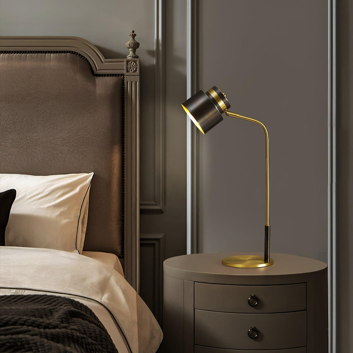A Clérachic bedside lamp on a round taupe cabinet with dark knobs, set against a padded headboard with a carved finial.