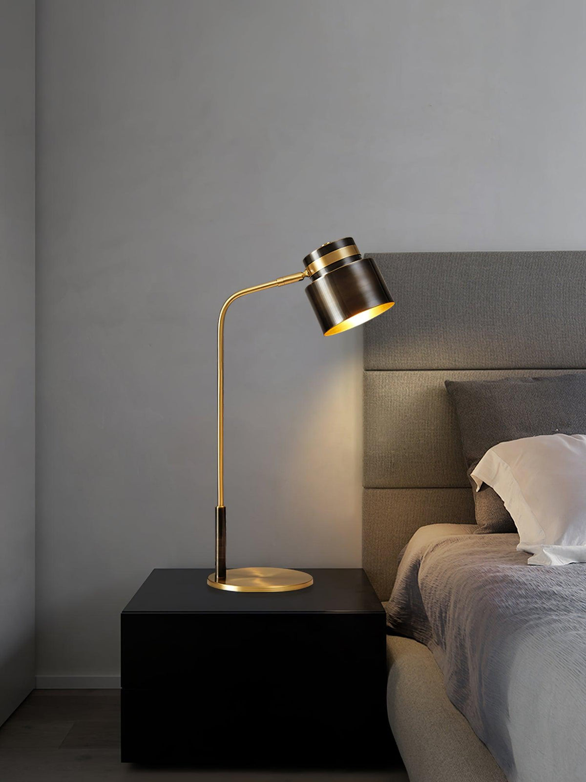 A Clérachic table lamp lit on a black bedside block, with a grey upholstered headboard and bedding beside it.