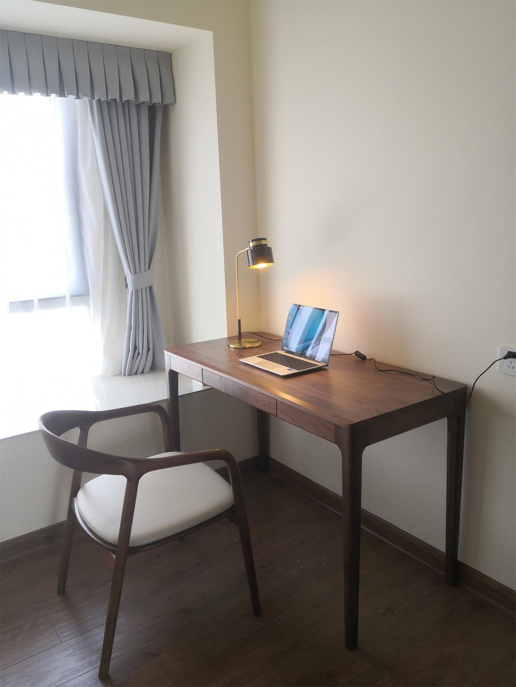 A small Clérachic lamp lit on a dark wood desk by a window with grey curtains, with a laptop and a pale cushioned chair.
