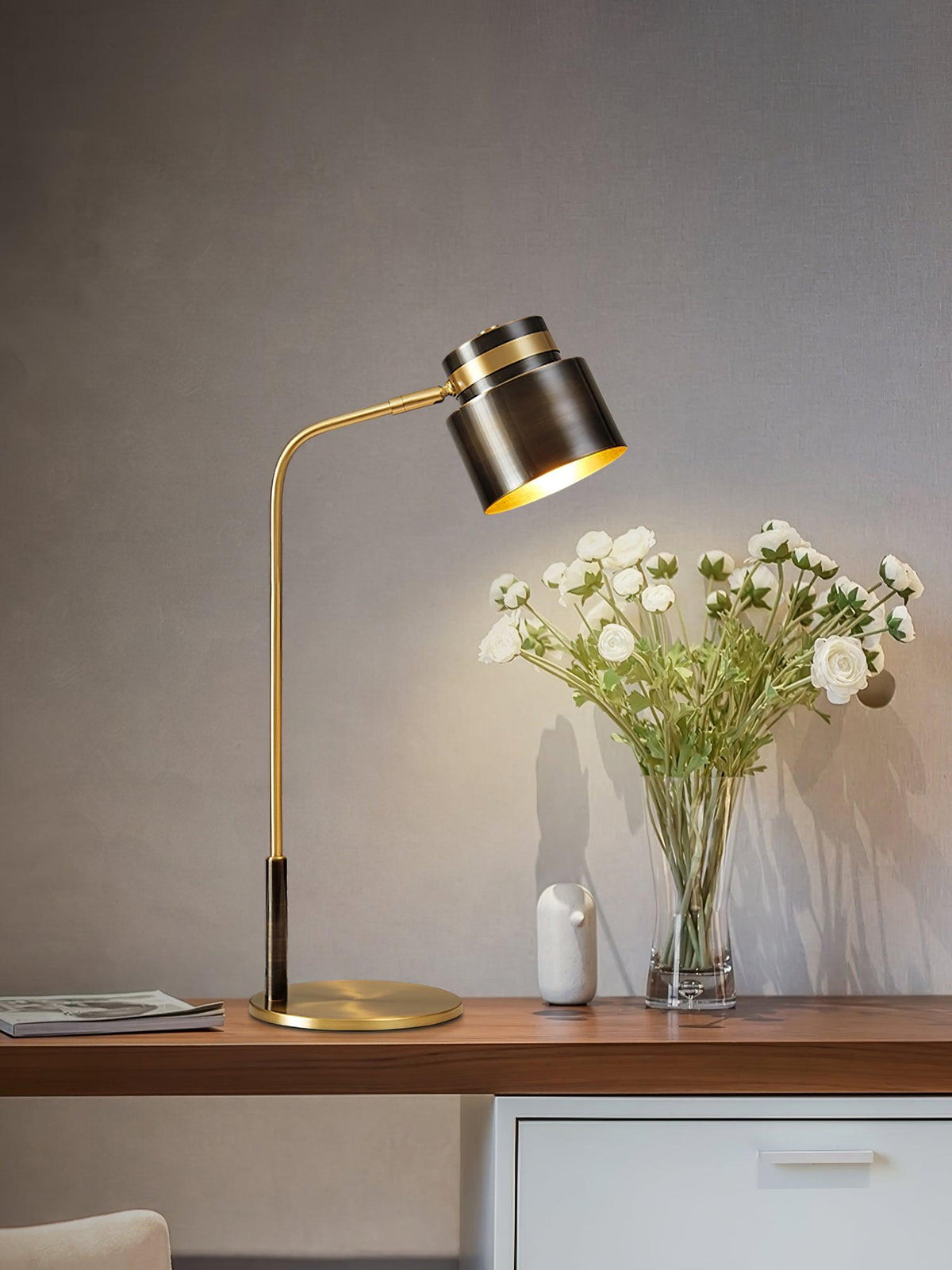 A Clérachic table lamp on a wooden shelf desk next to a glass vase of white flowers, with a magazine to the left.