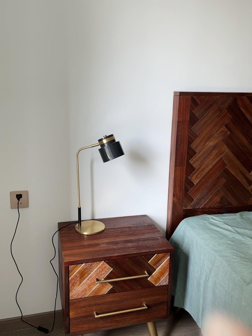 A Clérachic lamp on a herringbone-fronted wooden bedside table, with its black cord running down to a wall socket.