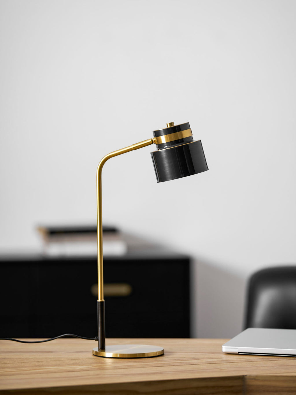 A Clérachic table lamp on a light wood desk, with a black chest of drawers and a laptop softly blurred behind it.