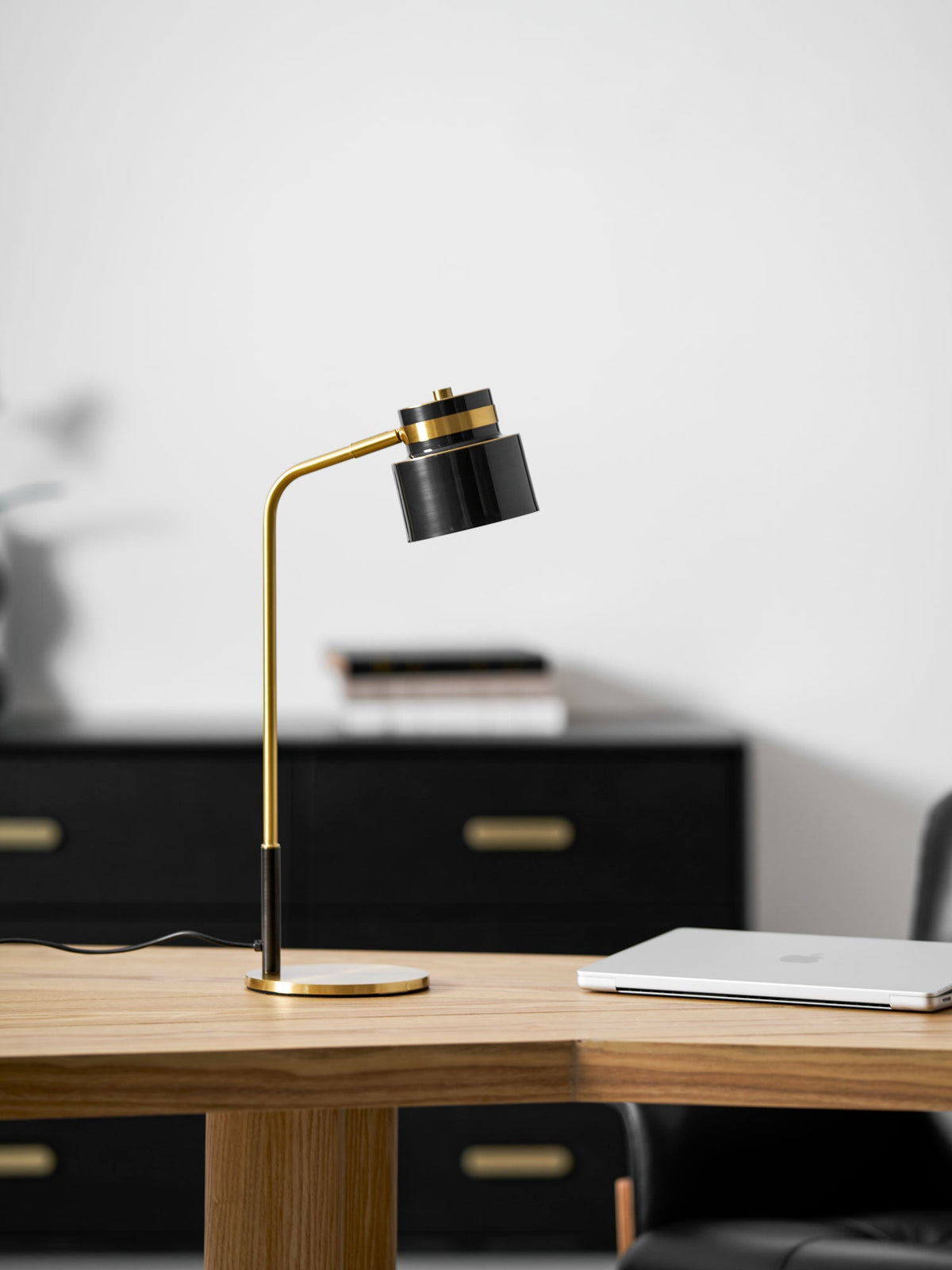 The Clérachic lamp on the corner of an oak desk, with a black sideboard and a closed laptop out of focus behind.