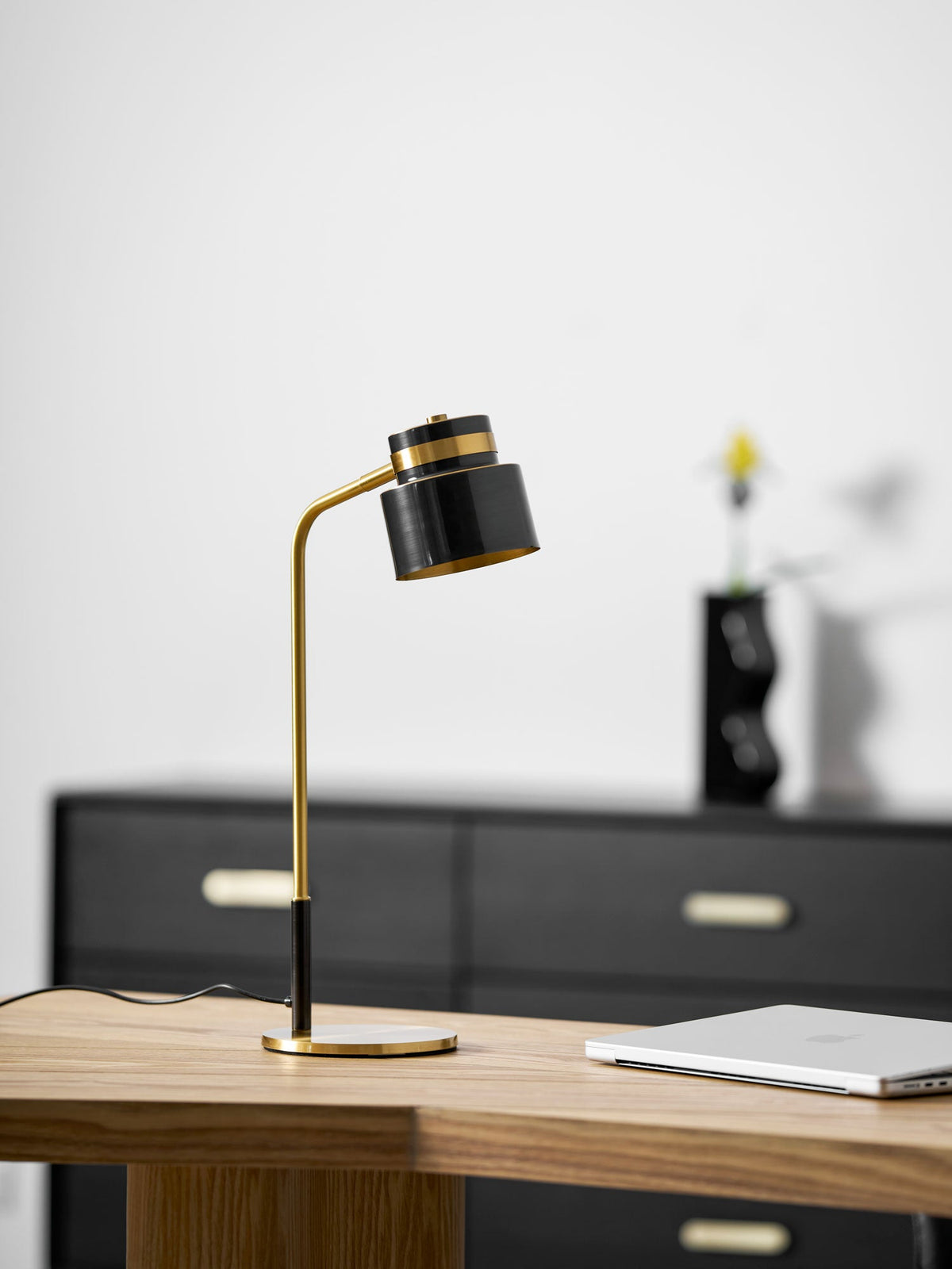 A Clérachic desk lamp on a wooden desk in front of a black sideboard with a small vase and yellow flower.