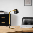 A Clérachic table lamp on an oak desk beside a silver laptop, with a black leather chair and framed print behind.