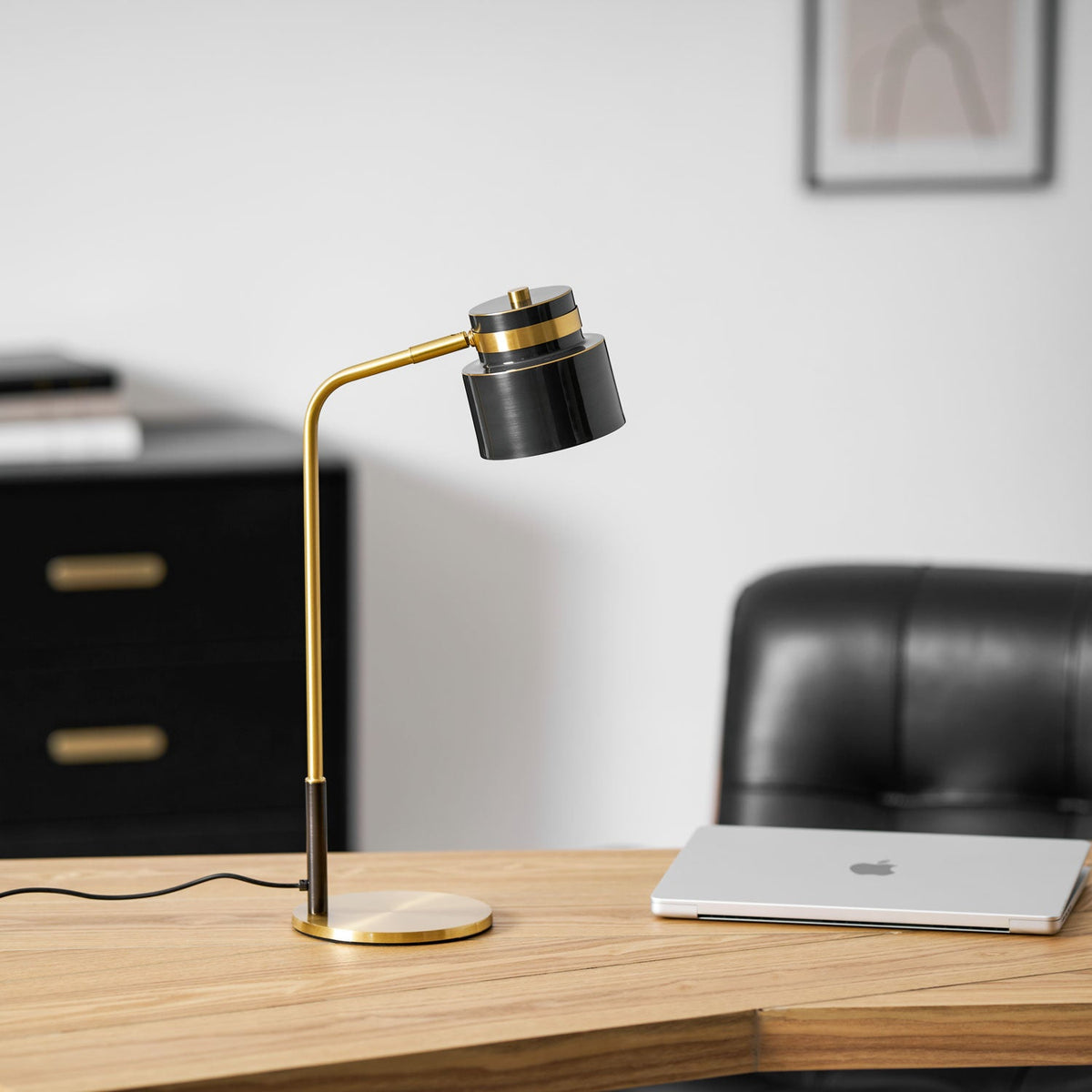 A Clérachic table lamp on an oak desk beside a silver laptop, with a black leather chair and framed print behind.