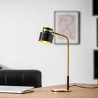 A Clérachic table lamp with its gold-lined shade tilted toward the camera, on a desk before a framed line drawing.